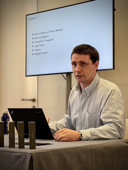 A man speaks at a table with a Lenovo laptop and small devices beside him, while a presentation slide lists “Contents,” including “Bitcoin Nodes and Bitcoin Network,” “Block Propagation,” “Transaction Propagation,” “Light Clients,” “Indexers,” and “Regtest Example.”