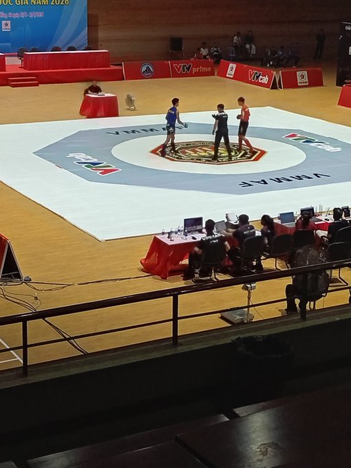 Two martial arts competitors stand on an octagonal mat in a gymnasium, with banners reading "VMF" and "Vinarme" visible around the perimeter during what appears to be a Vovinam tournament.