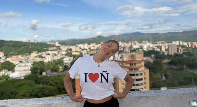 Woman wearing a white 'I ❤ Ñ' t-shirt posing outdoors with a cityscape and mountains in the background.