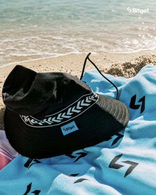 A black baseball cap and a light blue towel, both branded with the 'Bitget' logo, rest on sand at the edge of the ocean.