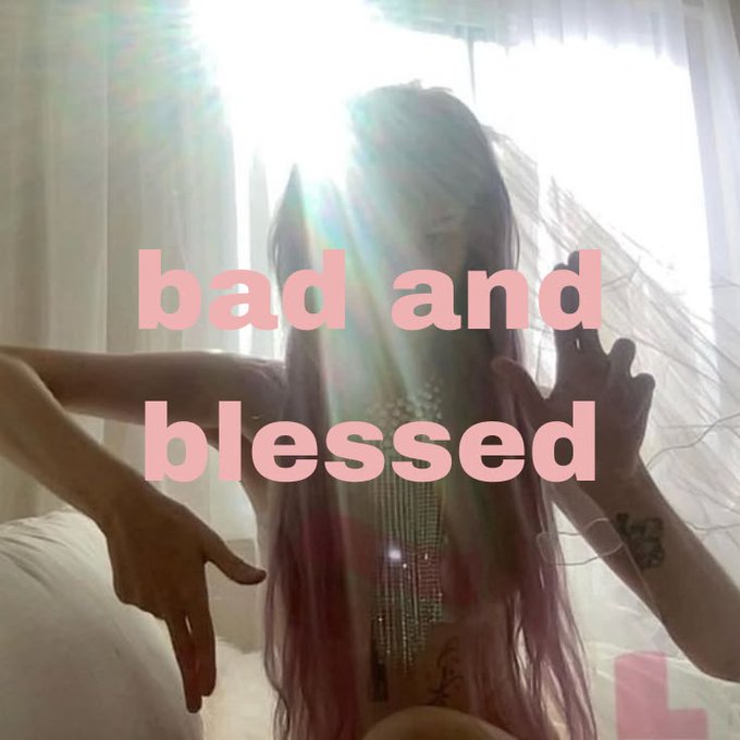 A person with long hair poses with hands raised in front of bright backlit curtains, with the pink text "bad and blessed" overlaid across the image.