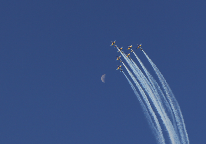 A formation of six jets flies through a clear blue sky, leaving white smoke trails behind them as a crescent moon is visible in the distance.