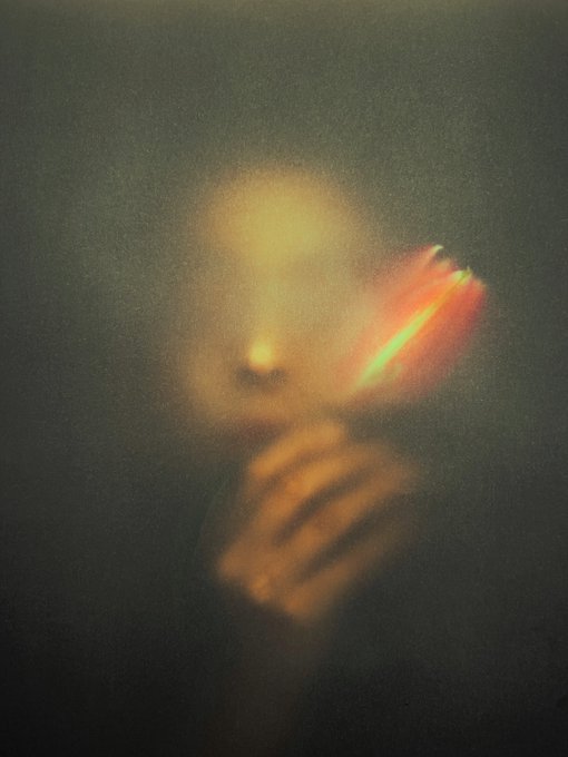 A dark, blurred photograph featuring a person holding a glowing object near their face, with warm orange and yellow light illuminating the scene against a black background.