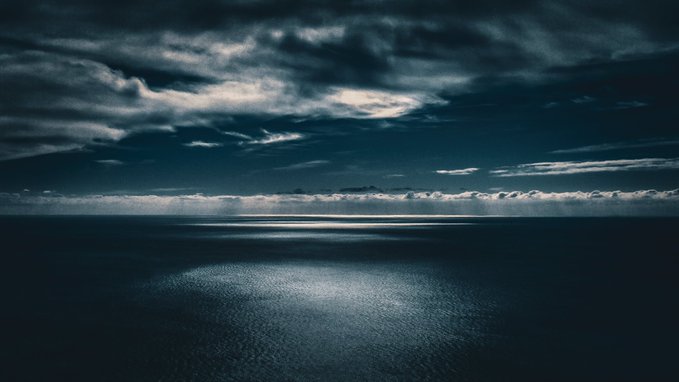 The image depicts a serene scene of an ocean or sea with a small island in the distance, under a cloudy sky. The vast body of water appears deep blue and still, creating a peaceful atmosphere. The island is relatively small, contrasting with the expansive nature of the surrounding area. There are no discernible texts or human-made objects in the image; it's a tranquil visual of natural beauty.