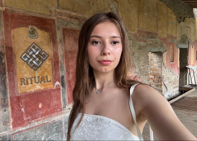 A young woman taking a selfie in front of an ancient Roman wall with red and yellow frescoes, one panel displaying a Celtic knot symbol with the word 'RITUAL'.