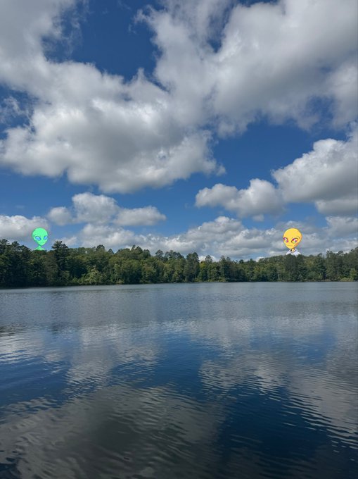 A serene lake is shown under a blue sky with white clouds, featuring a green alien emoji on the left and a yellow face emoji on the right hovering over the water.