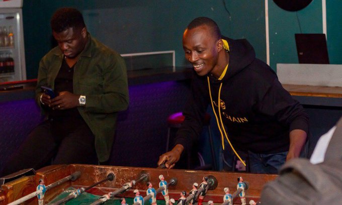 Two men sitting at a foosball table, one in a green jacket looking at his phone and the other in a black hoodie smiling while playing.