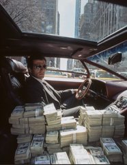 A man in a suit and sunglasses sitting in a luxury car surrounded by stacks of cash.