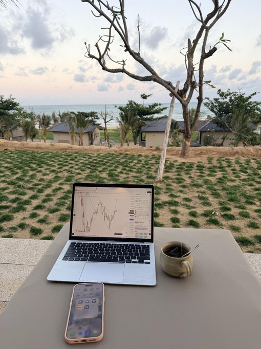A laptop displaying a stock chart sits on a table outdoors overlooking a grassy area and the sea, accompanied by a smartphone and a coffee mug.
