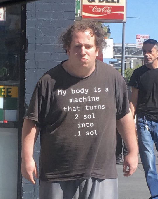 A man with a disheveled appearance stands on a city street, wearing a black t-shirt that reads "My body is a machine that turns 2 sol into .1 sol," capturing the relatable, humorous spirit of the "legend in the wild" reference.