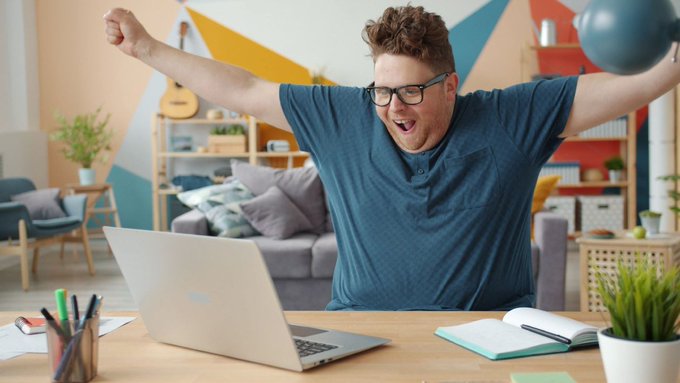 A young man with glasses celebrates with raised arms in front of a laptop in a colorful, modern living space.