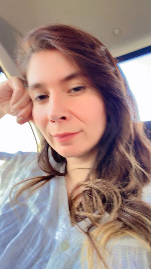 A selfie of a young woman with long brown hair wearing a light blue striped shirt, resting her head on her hand while sitting inside a vehicle.