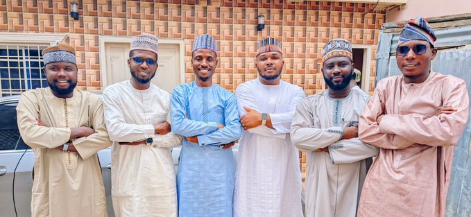 Six men wearing traditional Nigerian attire and caps posing together with their arms crossed in front of a brick building.