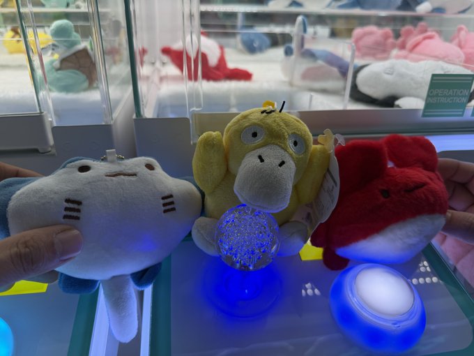 A hand holds two small plush toys—one white and one red—next to a yellow Psyduck plush sitting on an illuminated arcade machine, with other plush toys visible in the background case.