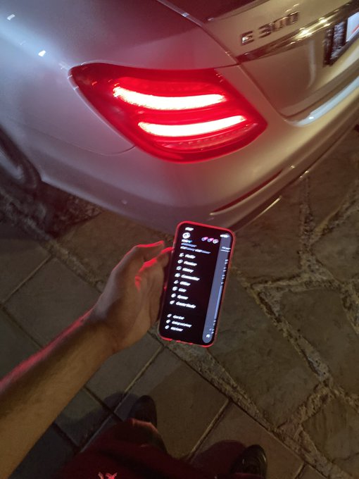 A person holding up a smartphone displaying a chat interface in front of the rear of a white Mercedes-Benz E 300 with illuminated red taillights.