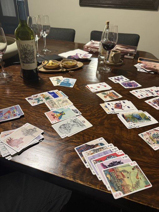 A wooden dining table set up for a tarot card reading session, featuring multiple tarot cards spread across the surface, a bottle of Amador wine, and wine glasses.