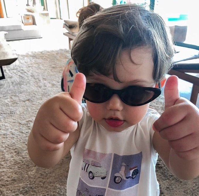 A close-up photo of a toddler wearing oversized black sunglasses and giving two thumbs up, dressed in a white shirt with a vehicle print.