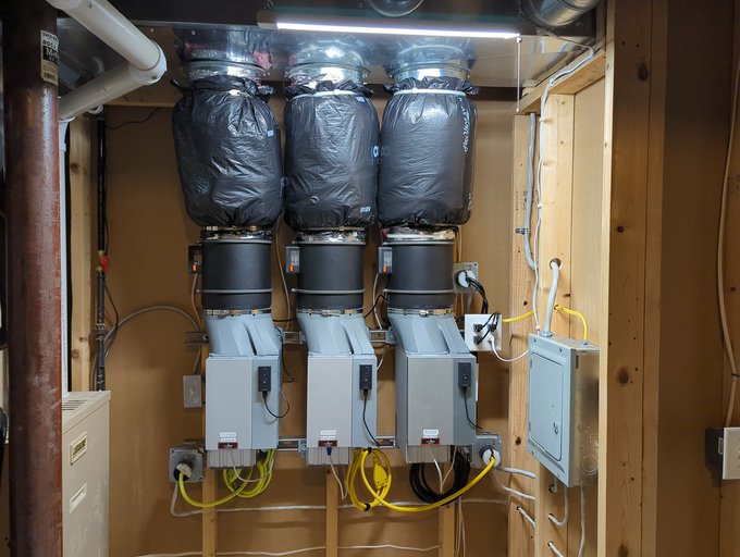 The image shows a room with three large air conditioning units mounted on the wall, each covered by black plastic bags. There are also several wires visible in the scene, indicating that these units may be part of a complex system for Bitcoin mining or cryptocurrency trading. The presence of multiple Bitcoin mining computers suggests that this space is likely used as a workspace for such activities.