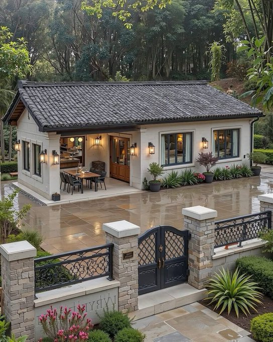 A modern, single-story house with a dark tiled roof, white walls, and an inviting outdoor patio area, surrounded by a stone wall and decorative iron gate, set in a lush, green environment.
