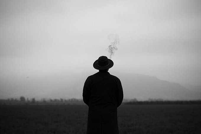 A black-and-white photograph of a person in a hat and long coat seen from behind, smoking a cigarette in an open field under a hazy sky.