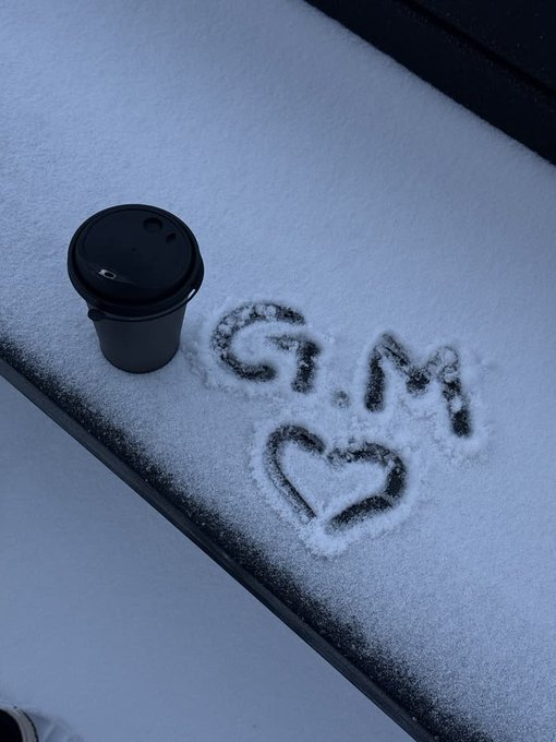 A black coffee cup sits on a snow-covered surface next to the initials 'G.M' and a heart drawn into the snow.