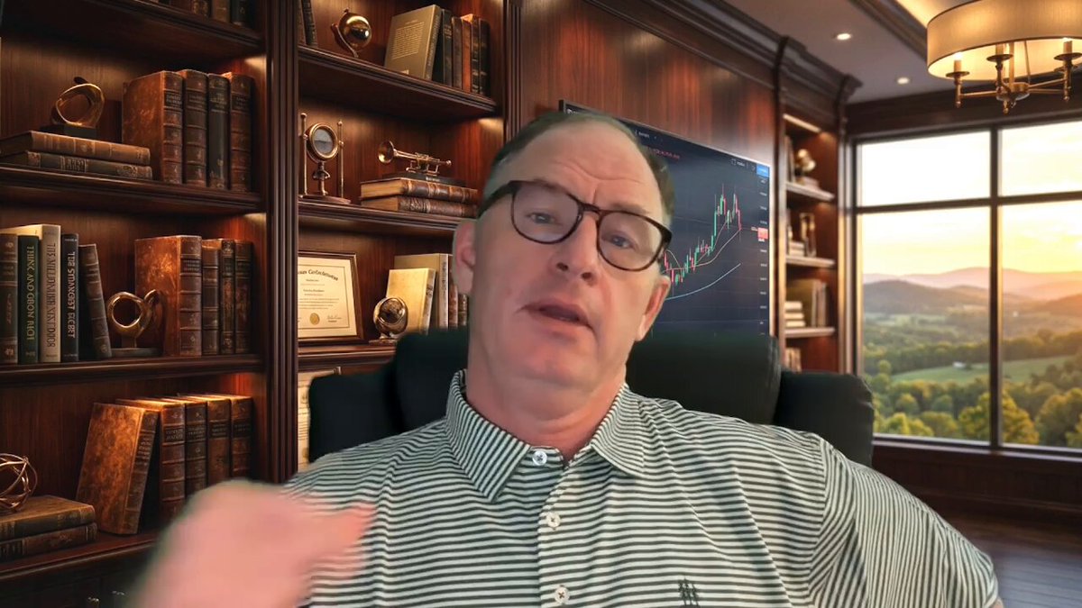 The image features a man sitting in an office chair, wearing glasses and a striped shirt. He appears to be giving a presentation or participating in a video call. The room has a window that offers a view of mountains, suggesting a scenic location for the meeting. There are several bookshelves filled with various books around the room, indicating a well-read environment.