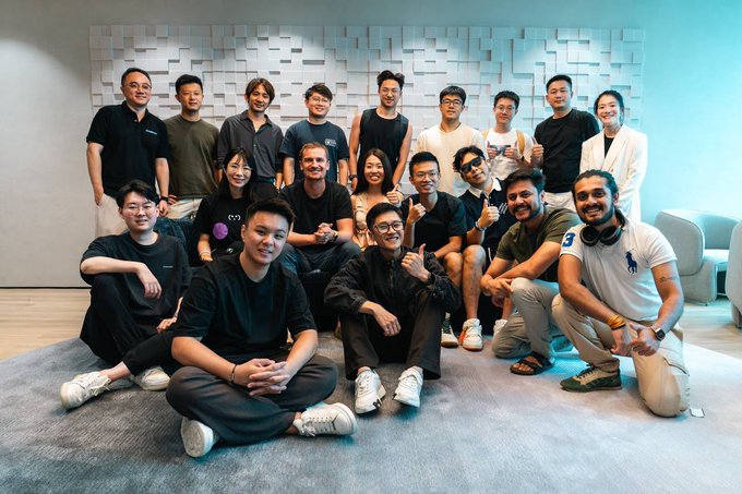 A group photo of approximately 17 people posing together in a modern room with textured white walls, smiling and making casual gestures for the camera.