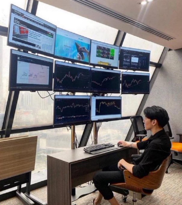 A man sits at a desk in a high-rise office analyzing multiple stock charts and financial data displayed across a large multi-monitor setup.