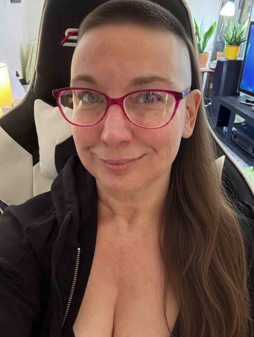 The image features a woman with long brown hair wearing glasses, smiling at the camera. She has short hair that is styled into a ponytail. The background of the photo shows a chair and a TV screen, suggesting that she might be sitting in front of these items while posing for the picture. The caption "Mulletized" appears to be related to her hairstyle or appearance, possibly indicating that she has recently undergone a haircut or a change in her hair style.