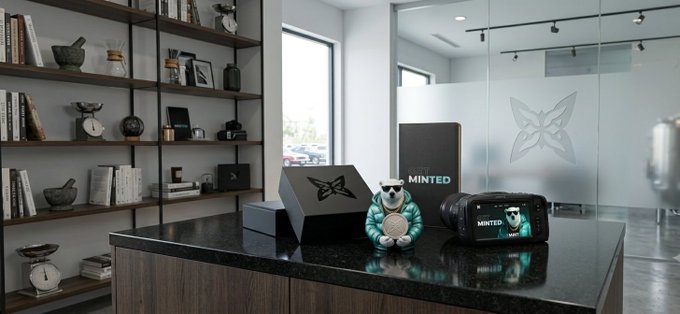 Display on a dark counter featuring a small bear figurine in a teal jacket alongside a black box and case, both bearing a butterfly logo and the word 'MINTED'.