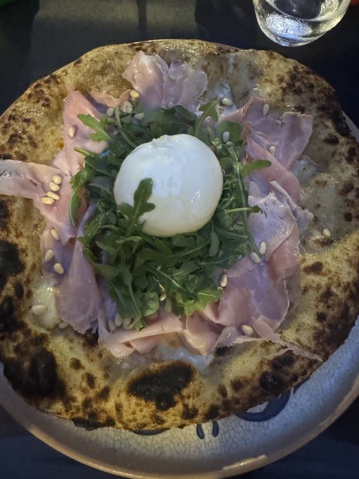 A flatbread pizza topped with prosciutto, arugula, pine nuts, and a large ball of burrata cheese in the center, served on a patterned plate.