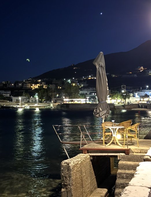 The image features a beautiful view of a waterfront at night, with a boat docked nearby. There are two chairs positioned near the water's edge, providing an ideal spot for relaxation or contemplation. A large umbrella stands over one of the chairs, offering shade from the moonlight and enhancing the ambiance of the scene. The backdrop is a picturesque view of a mountain range under a dark sky, adding to the overall tranquility of the setting. In the image, there are also two small boats visible in the water near the dock.