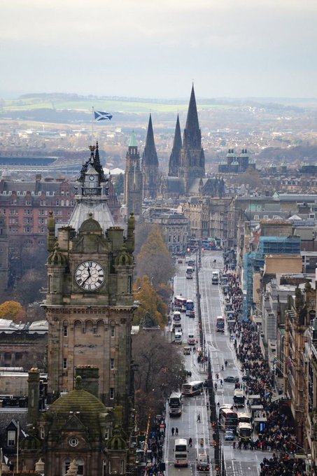 An elevated view of Edinburgh, Scotland, featuring the Balmoral Hotel clock tower in the foreground, a long street with traffic and pedestrians, and a Scottish flag flying.