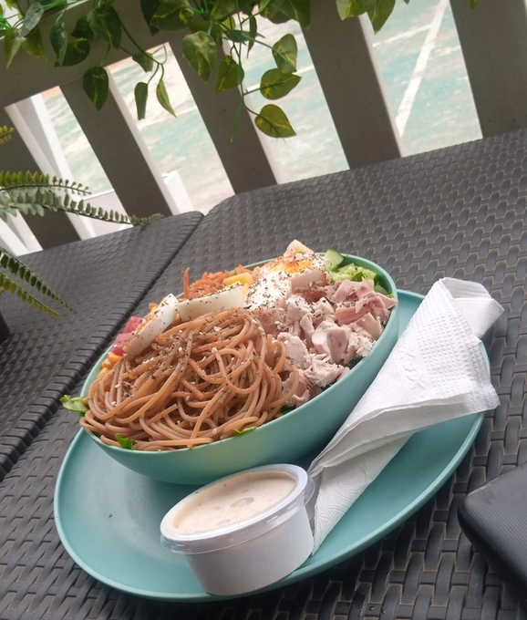 A teal bowl filled with soba noodles, sliced meat, and vegetables sits on a matching plate on a black woven table, with a small container of sauce and a napkin beside it.