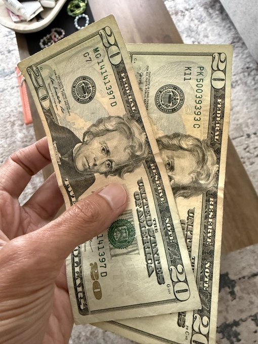 A hand holding two slightly worn $20 U.S. bills (serial numbers PK50039393 C and ML14113970 G) in front of a marble surface with scattered jewelry visible in the background.