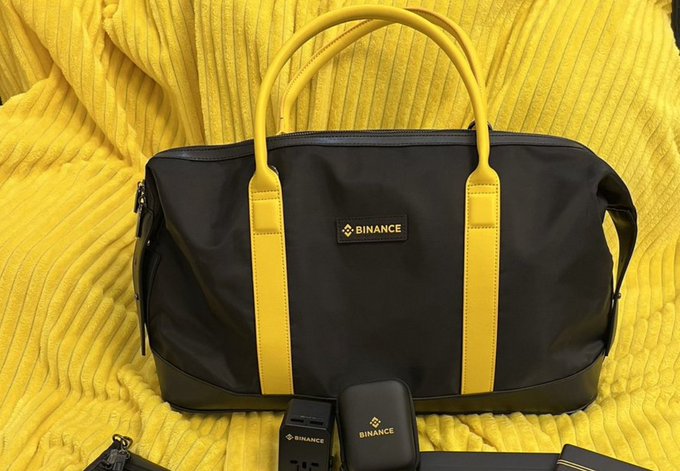 A black and yellow duffel bag branded with the Binance logo sits on a yellow ribbed sweater, accompanied by Binance-branded tech accessories.