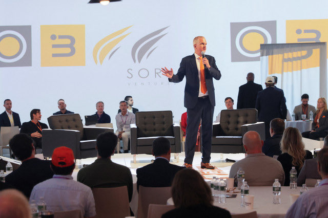 A man in a dark suit and orange tie speaks into a microphone on stage at a 'SOR... VENTURE' event, with an audience seated in front of him.