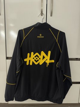 A black jacket hanging on a wall, featuring yellow piping and a large yellow graffiti-style logo on the back with a small "BINANCE" text near the collar.