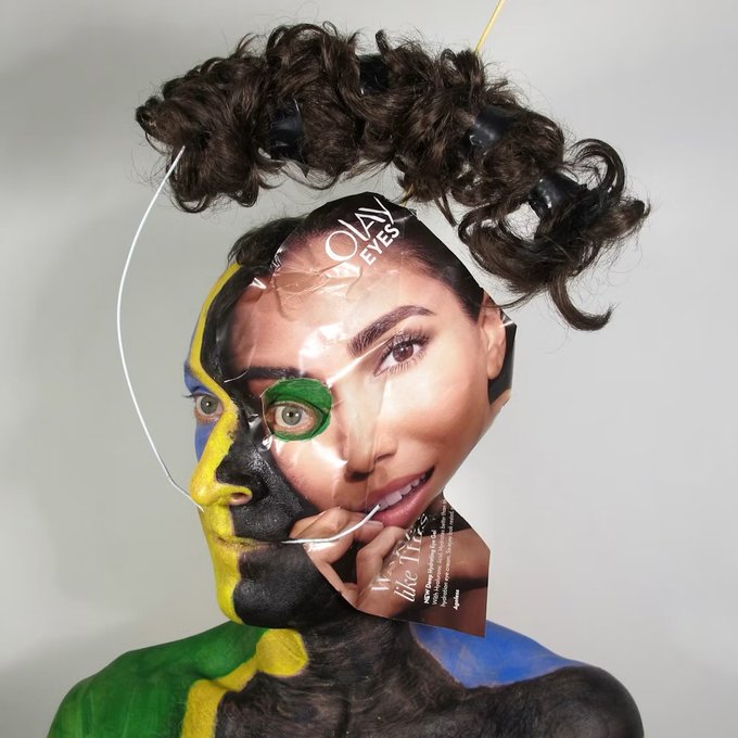A striking portrait of a person with curly hair, wearing a mask that is half colorful paint and half a torn magazine cover showing a face with green eyeshadow and the text 'OLAY EYES'.