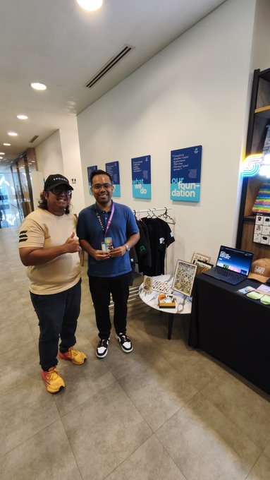 Two men posing in what appears to be a community space, with one giving a thumbs up while holding a drink, near a table displaying merchandise and a laptop under 'Our Foundation' signage.