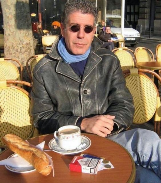 The image shows an older man sitting at a table with a cup of coffee and a sandwich. The man has sunglasses on his head and appears to be enjoying his meal outside, possibly in a cafe or restaurant setting. There are other people present in the scene as well, indicating that this is a public space where multiple individuals can enjoy their time.