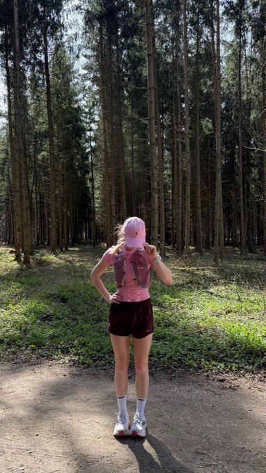 A person wearing a pink cap, pink top, and maroon shorts stands on a dirt path in a dense pine forest, with their face obscured.