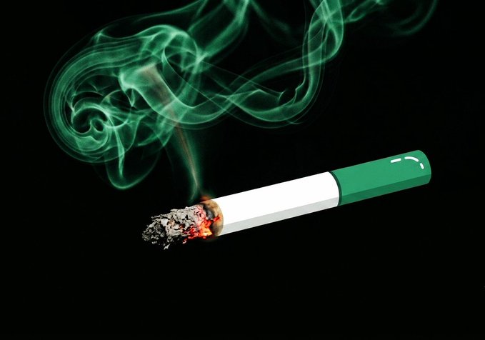 A lit cigarette with a green filter burns against a black background, with the ember glowing orange and red ash crumbling away as green-tinted smoke swirls upward.