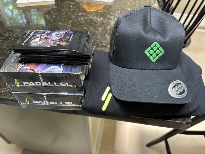 A stack of 'Parallel' trading card boxes sits next to a black and green snapback hat on a dark granite countertop.