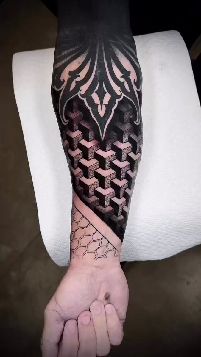 The image features a person's arm with an intricate black tattoo design, including geometric shapes like squares and diamonds. The tattoo appears to be on the inner side of the forearm or wrist area. The background of the photo has a white towel hanging from a hook, which contrasts nicely with the dark tones of the tattoo. The caption of the image reads "Today's efforts. Wish they were hexagons... but still pretty cool @RichardHeartWin #PulseChain".