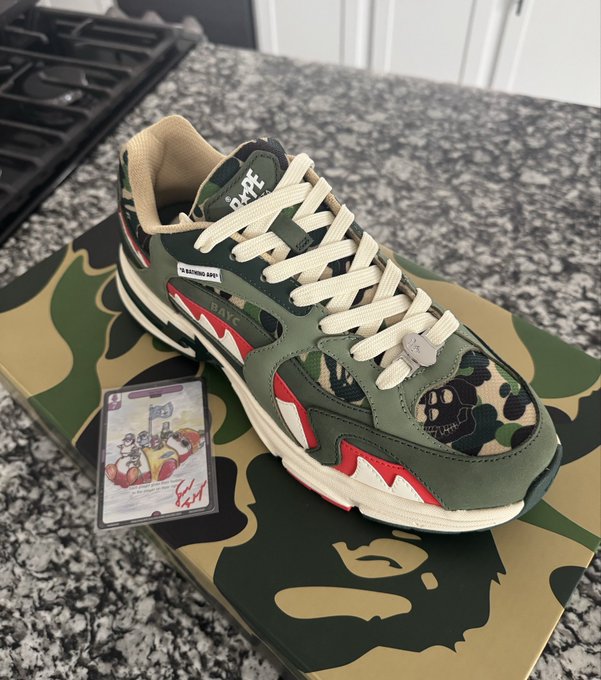 A pair of green, beige, and red camouflage BAPE sneakers with shark teeth design rests on its matching camouflage shoebox, accompanied by a small collectible card on a granite countertop.