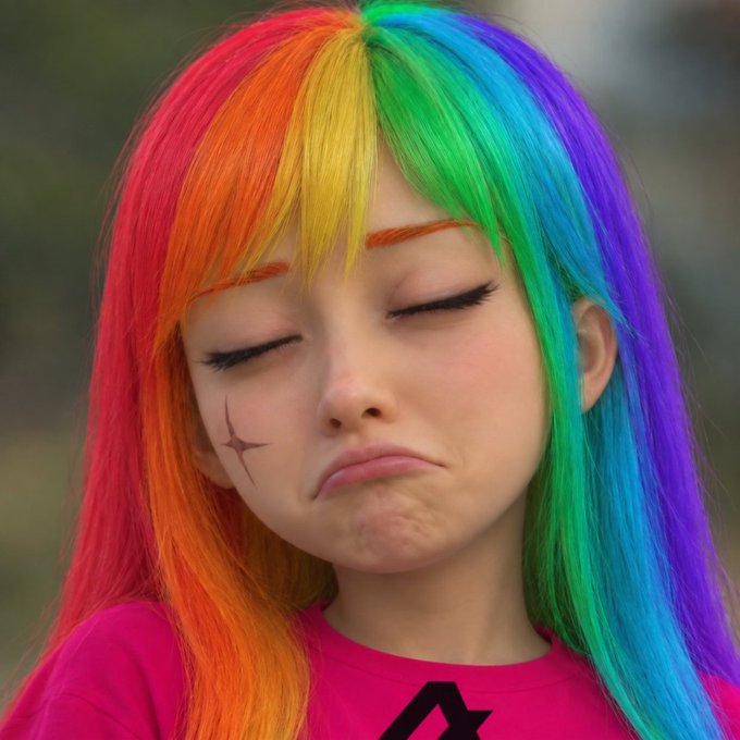 A woman with vibrant rainbow-colored hair and orange eyebrows makes a pouting expression, wearing a pink shirt.