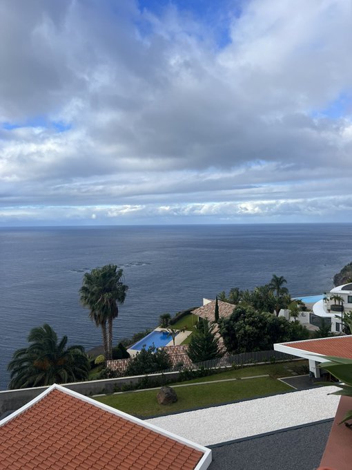 An ocean view from an elevated patio showing a cloudy sky, the sea, palm trees, and nearby residential buildings with orange tile roofs.
