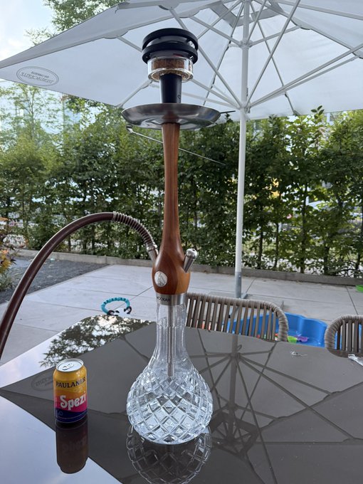 An outdoor patio scene featuring a tall glass and wood hookah on a dark reflective table next to a can of Paulaner Spezi soda, set under a large white umbrella.