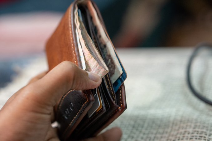 A hand opens a brown leather bifold wallet stuffed with folded paper currency and several plastic cards, with the embossed letters "me" visible on the front leather, visually echoing the accompanying post's self-deprecating message about Zerion—a crypto portfolio tracker app—showing gains on other wallets while the user's own "lifeless" portfolio fails to perform, the physical full wallet ironically contrasting with the implied financial frustration expressed in the text.